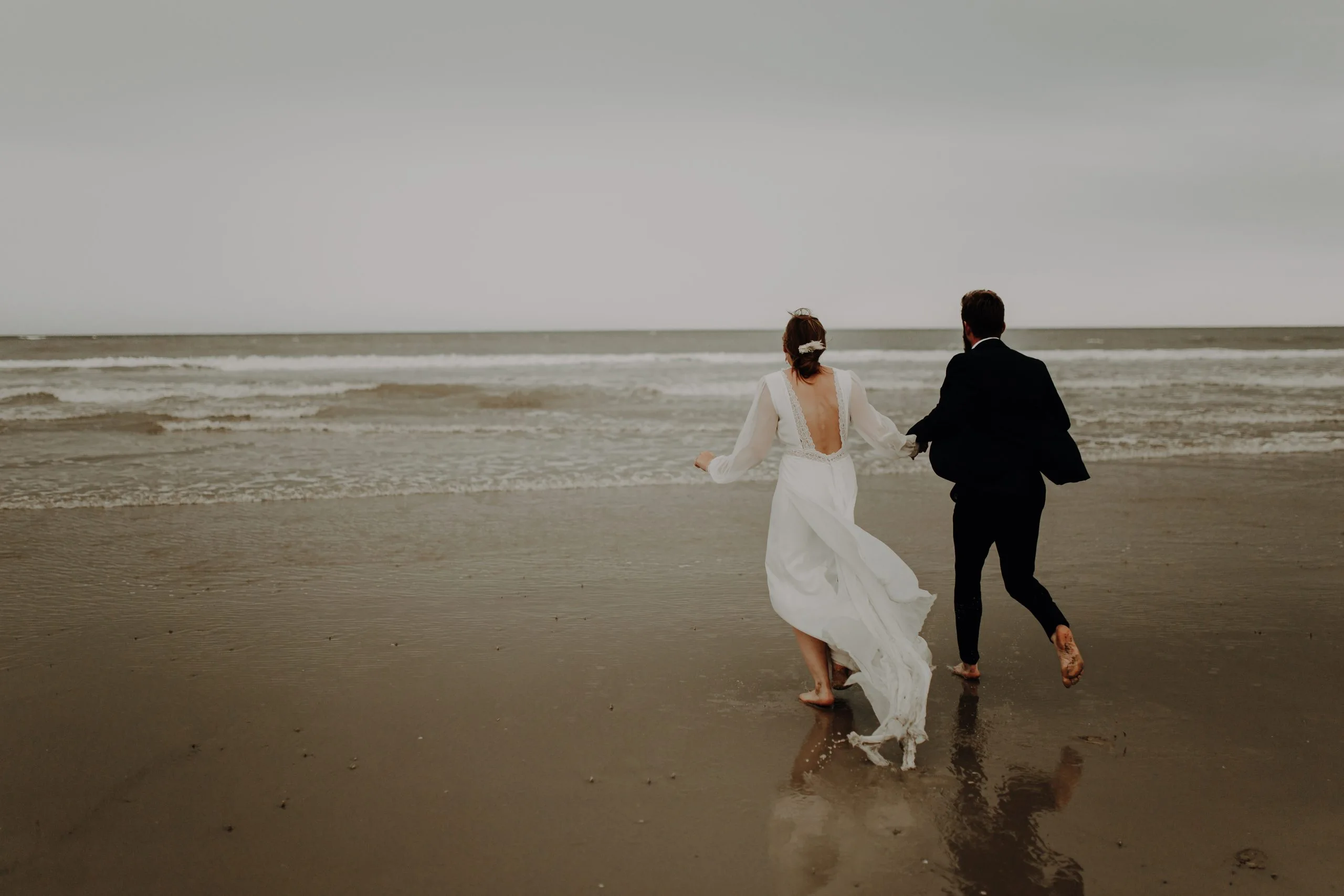 Blanche et Lucas courent main dans la main vers la mer sur la plage du Touquet, pieds nus dans le sable mouillé et vêtus de leurs tenues de mariage.