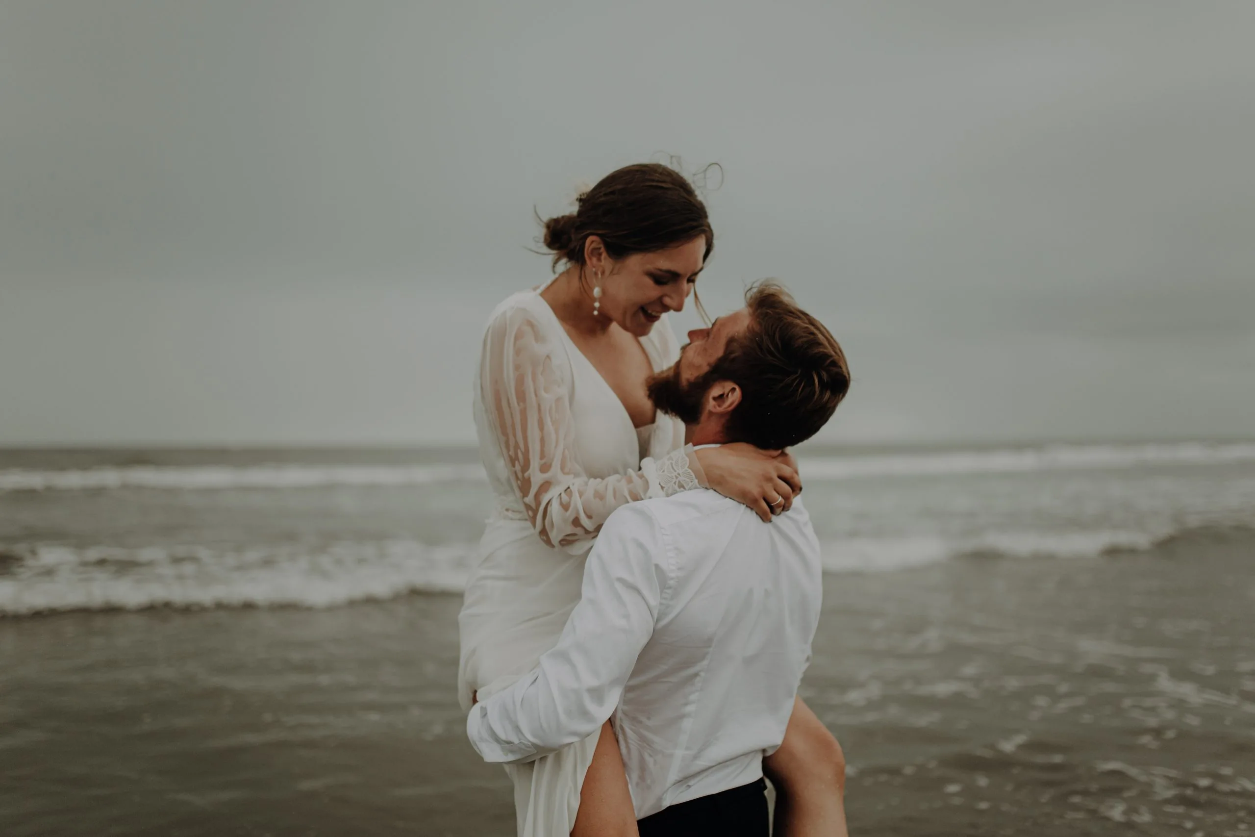 Blanche et Lucas assis face à face sur le sable humide du Touquet, dans leurs tenues de mariage, sous un vaste ciel gris.