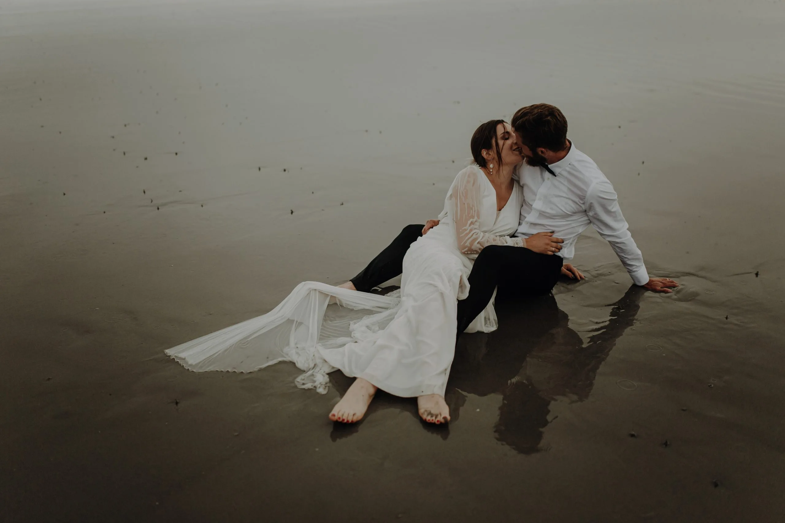 Blanche et Lucas s'embrassent assis sur le sable mouillé de la plage du Touquet, avec la longue traîne de la robe de mariée étendue derrière eux.