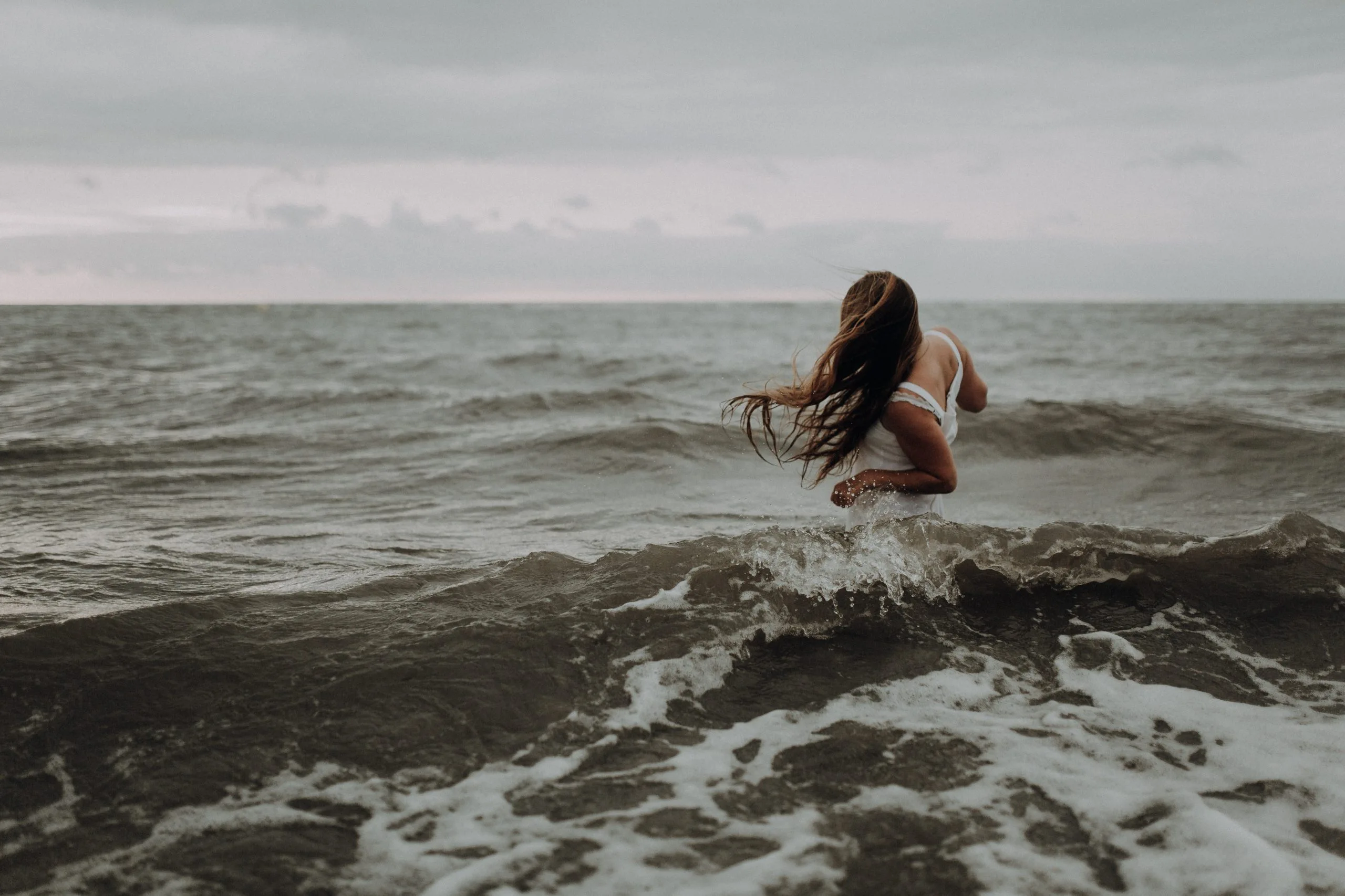 Femme en robe blanche entrant dans les vagues de la mer sous un ciel couvert