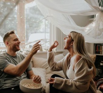 Couple jouant avec des oreillers et des draps blancs dans une chambre lumineuse.