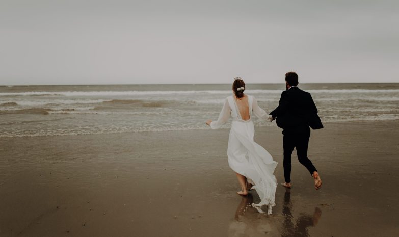 Blanche et Lucas courent main dans la main vers la mer sur la plage du Touquet, pieds nus dans le sable mouillé et vêtus de leurs tenues de mariage.