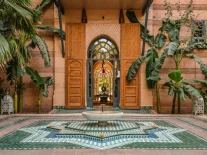 Entrée majestueuse d’un riad à Marrakech avec porte marocaine sculptée, fontaine centrale et sol en zellige vert et blanc, entourés de végétation tropicale.