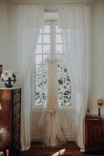 Robe de mariée en dentelle ivoire suspendue devant une grande fenêtre à petits carreaux, encadrée de rideaux blancs, dans une chambre du château.