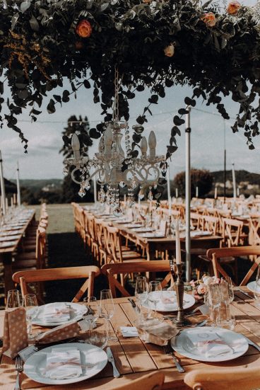 Gros plan sur la décoration des tables de mariage avec chandeliers suspendus et végétation en extérieur.