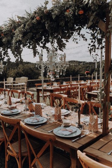 Gros plan sur la décoration des tables de mariage avec chandeliers suspendus et végétation en extérieur.