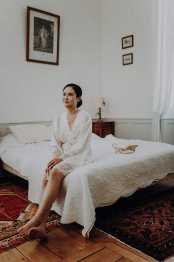 eune mariée assise sur un lit dans une chambre lumineuse du château, vêtue d'un peignoir blanc.