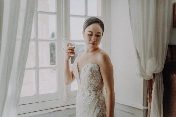 Portrait de la mariée en robe de dentelle vaporisant son parfum devant une fenêtre aux rideaux blancs pendant ses préparatifs.
