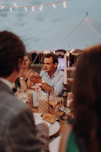 Invités réunis autour des tables dressées pendant une réception de mariage en extérieur à la tombée du jour.