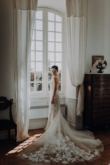 Mariée en robe blanche photographiée près d'une grande fenêtre aux rideaux clairs dans une chambre du Château Lacoste.