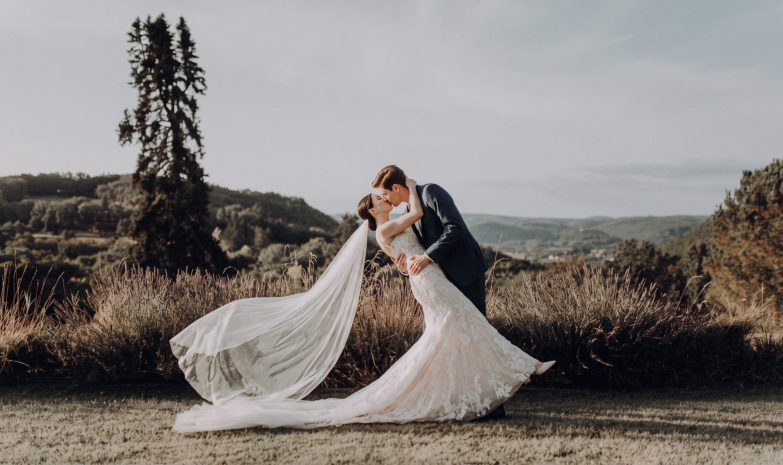 Couple de mariés s'embrassant dans un paysage naturel avec le long voile de la mariée déployé derrière elle.