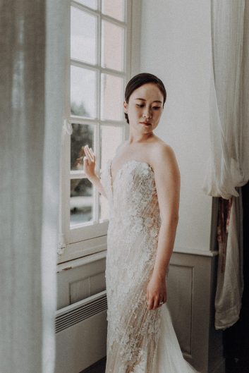 Portrait de la mariée portant une robe en dentelle près d'une grande fenêtre lumineuse du château.