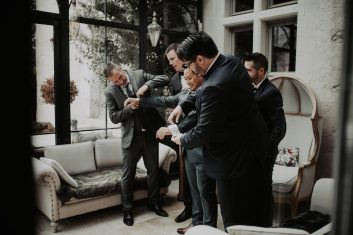 Château des Girards - Préparatifs entre amis Plusieurs hommes en costumes élégants discutant dans la cour du château pendant les préparatifs du mariage.
