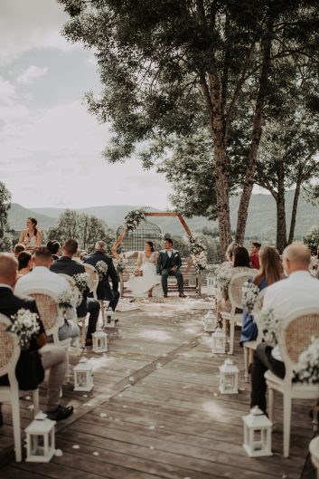 Château des Girards - Une cérémonie au jardin Vue d'ensemble d'une cérémonie de mariage en extérieur avec des invités assis sous les arbres et une arche fleurie.