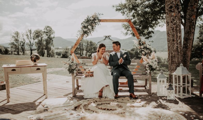 Couple de mariés se tenant devant une arche fleurie lors de leur cérémonie de mariage en extérieur.